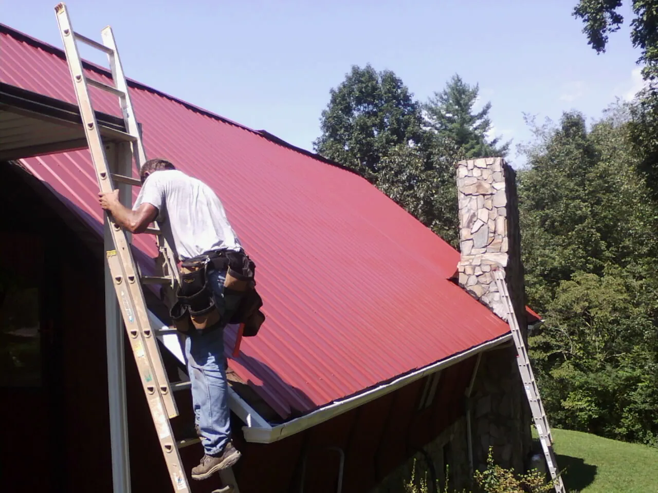 Roofer on a ladder beside a red metal roof and stone chimney — Soffit & Fascia Repair by Roofing Brothers in Slippery Rock